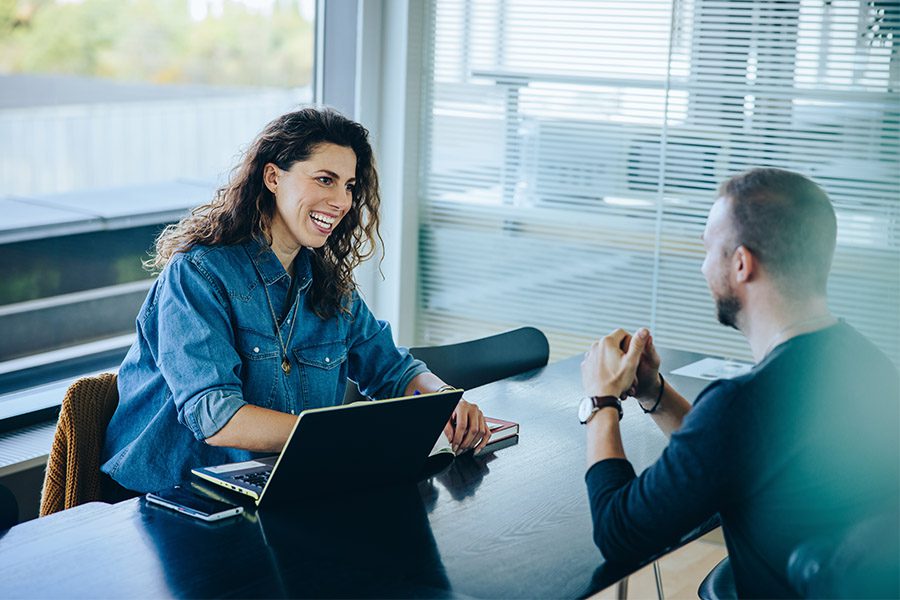 HR & Payroll Solution - Woman Smiling and Talking to a Man While Sitting at a Black Desk With Her Laptop Open in the Office on a Nice Day