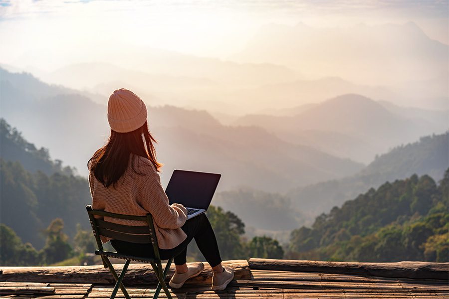 Blog - Woman Sitting Out on a Wooden Deck Over Looking a Beautiful View of Moutains As She Works on Her Laptop