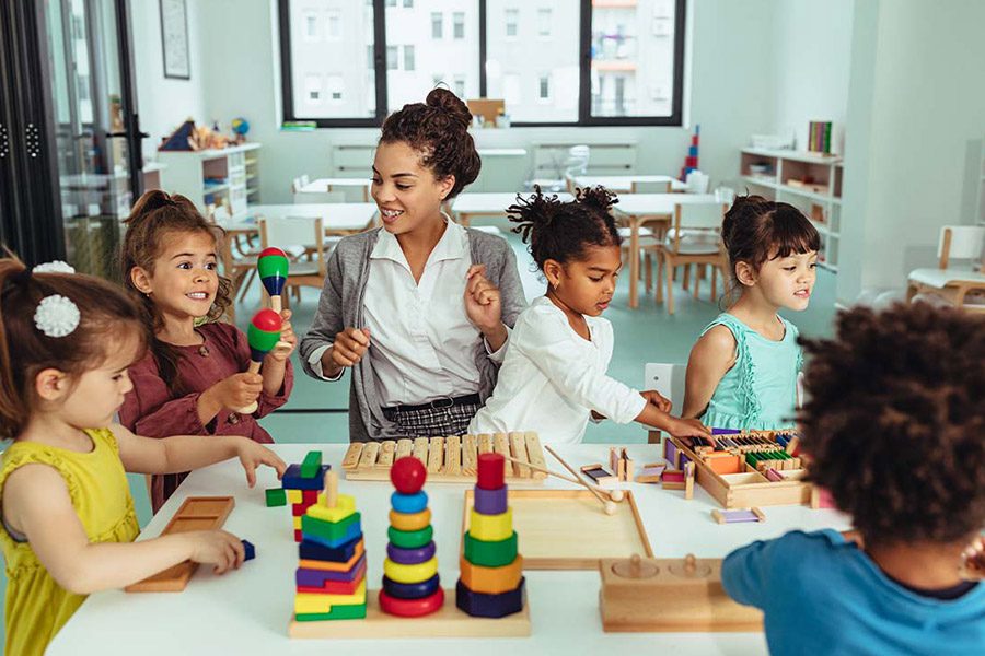 Child Care Center Insurance - Day Care Employee or Kindergarten Teacher Playing with Young Children in the Classroom and Creating Artwork and Building Blocks Together