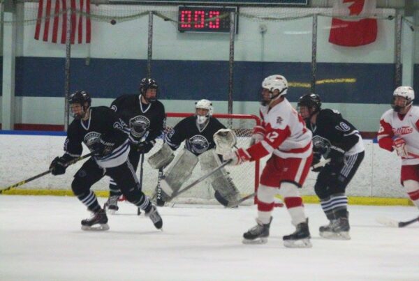 Hockey players in black and white jerseys skating against players in red and white jerseys during a competitive game, with a goalie in position in front of the net.